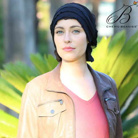 A woman in a black Angelle Chemo Beanies® scarf and a brown leather jacket stands outdoors with green plants behind her. The Chemo Beanies® logo appears in the top right corner.