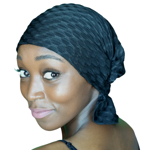 A woman with smooth brown skin and bright eyes smiles softly over her shoulder, wearing a black Chemo Beanies® Emma beanie and a matching top against a plain black background.