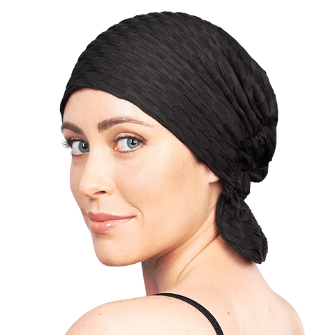 A fair-skinned, light-eyed woman wears the Chemo Beanies® Emma textured black head cover, tied at the back. She glances over her shoulder with a slight smile against a plain black background.