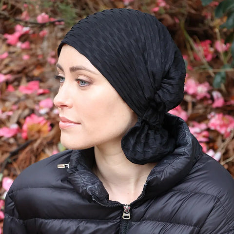 A woman wearing the Scarves - Chemo Beanies® - Emma by Chemo Beanies® and a black puffer jacket sits outdoors among fallen brown leaves and pink petals, looking to the left with a calm expression.
