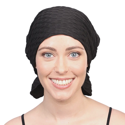 A smiling woman with bright blue eyes wears the Chemo Beanies® Emma chemo head cover in black, paired with a matching top. She is facing forward against a plain background.