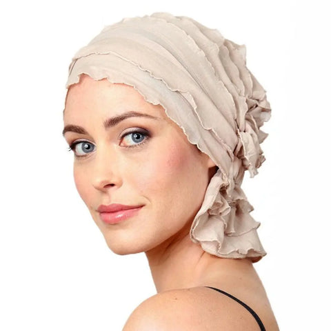 A woman with light skin and blue eyes smiles softly while wearing the Chemo Beanies® Megan ruffled beanie in light beige, by Chemo Beanies®, against a plain white background.