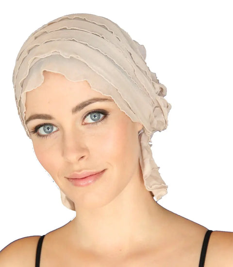 A light-skinned, blue-eyed woman smiles softly at the camera against a white background, wearing the Chemo Beanies® Megan scarf—a sheer, beige cancer headwear style with layered ruffled edges and a black strap top.