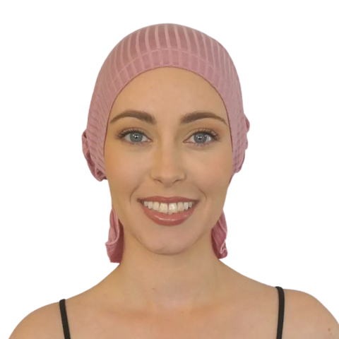 A woman with light skin and blue eyes smiles while wearing the Chemo Beanies® Taylor, a light pink pleated chemo beanie, paired with a black strap top against a plain background.