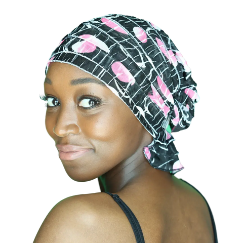 A woman with glowing skin smiles over her shoulder, wearing the Chemo Beanies® Delia—a black slip-on chemo beanie with a pink and white pattern—styled as elegant hair loss headwear, paired with a black tank top against a bright background.