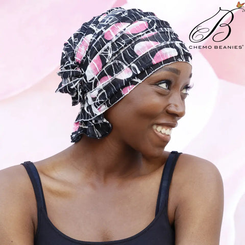 A smiling woman in a Chemo Beanies® Delia patterned slip-on beanie and black tank top looks to the side. The soft pink background features the Chemo Beanies® logo, highlighting comfortable hair loss headwear.