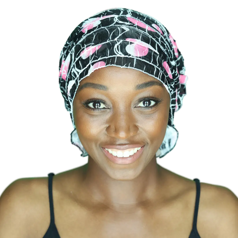 A smiling woman models the Chemo Beanies® Delia scarf—a stylish black and white chemo beanie with pink patterns—paired with a black spaghetti strap top, perfect for fashionable hair loss headwear.