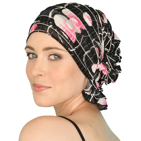 A woman with light skin wears the Chemo Beanies® Delia, a black floral chemo beanie by Chemo Beanies®, smiling softly over her shoulder against a plain white background.