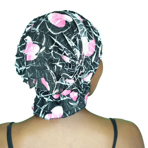 A person with medium brown skin, seen from behind in a black spaghetti strap top, wears the Delia Chemo Beanies® scarf by Chemo Beanies®, featuring a black and pink pattern.