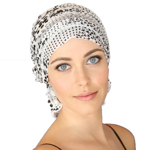 A woman with blue eyes and light makeup models the Chemo Beanies® Scarves - Reagan, a black and white printed chemo head cover, paired with a black-strapped top against a plain white background.