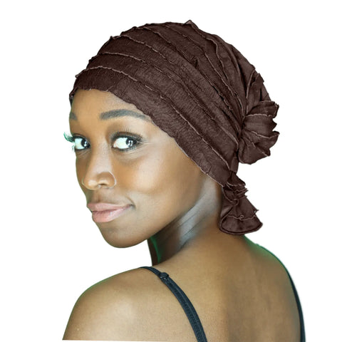 A woman with smooth brown skin and dramatic makeup looks over her shoulder, wearing the Chemo Beanies® Janice textured brown chemo head cover and a black strap top, set against a white background.