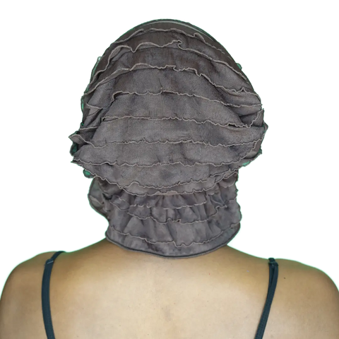 A person is seen from behind wearing the Chemo Beanies® Janice chemo beanie, featuring dark ruffles. Bare shoulders and thin black straps show against a plain black background.