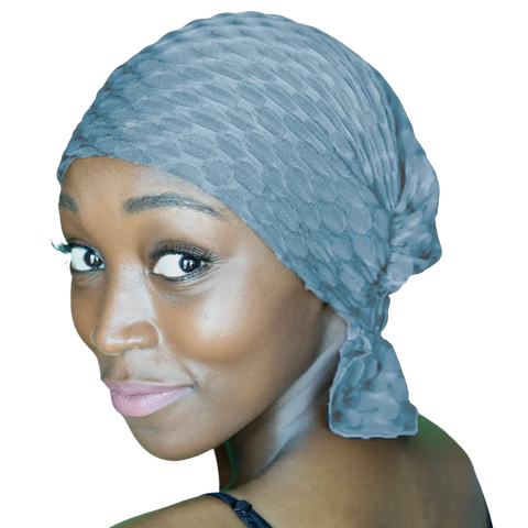 A woman with smooth brown skin smiles gently over her shoulder, wearing the Chemo Beanies® Zoe scarf in textured light blue, tied at the back. Her black strap top and a plain dark background highlight her expressive eyes.