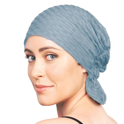A woman with light skin and blue eyes, dressed in black, smiles subtly as she looks over her shoulder wearing the Zoe Chemo Beanie® from Chemo Beanies®, a textured light blue scarf-style beanie tied at the back, against a white background.