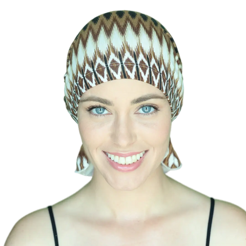 A smiling woman with bright blue eyes wears a patterned brown and white Chemo Beanies® Roberta scarf and a black spaghetti strap top against a plain black background.