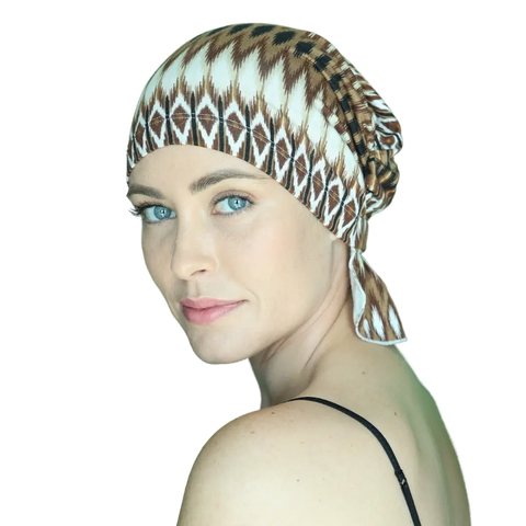 A woman with light skin and blue eyes models the Chemo Beanies® Roberta patterned chemo beanie, paired with a black spaghetti strap top, looking over her shoulder at the camera against a plain background.