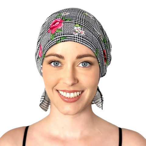 A smiling woman with light skin and blue eyes models the Chemo Beanies® Pixie—a black and white checkered chemo beanie with pink and red floral patterns—by Chemo Beanies®, photographed against a plain background.