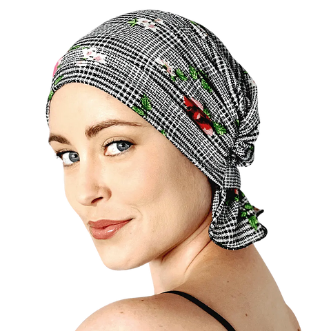 A light-skinned woman with blue eyes smiles softly, wearing a black-and-white floral Chemo Beanies® Pixie scarf and a black strap top, set against a black background.
