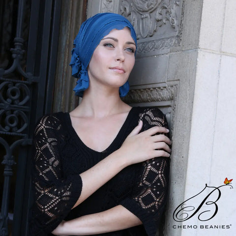 A woman wearing the Chemo Beanies® Muriel scarf and a black lace top leans against a stone wall, gazing thoughtfully into the distance. The Chemo Beanies® logo appears in the lower right corner.