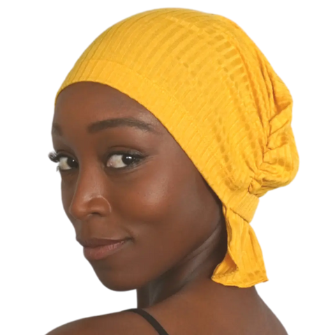 A woman with smooth dark skin wears the bright yellow Chemo Beanies® Martha chemo scarf, paired with a black strap top, looking over her shoulder at the camera with a gentle smile.