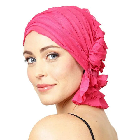 A woman with light skin and blue eyes models the Chemo Beanies® Carol chemo beanie in bright pink with ruffled details, smiling softly over her shoulder against a white background.