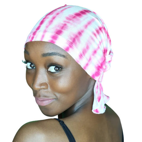 A woman with dark skin smiles confidently over her shoulder, wearing the Chemo Beanies® TANISHA CHEMOBEANIE® in pink and white tie-dye as cancer headwear. Her black strap top pops against the black background.