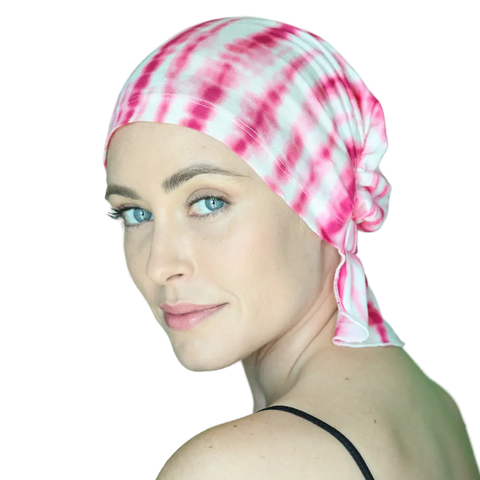 A light-skinned, blue-eyed woman in a black spaghetti strap top smiles softly over her shoulder while wearing the TANISHA CHEMOBEANIE® by Chemo Beanies®—a pink and white tie-dye cancer headwear—against a plain background.