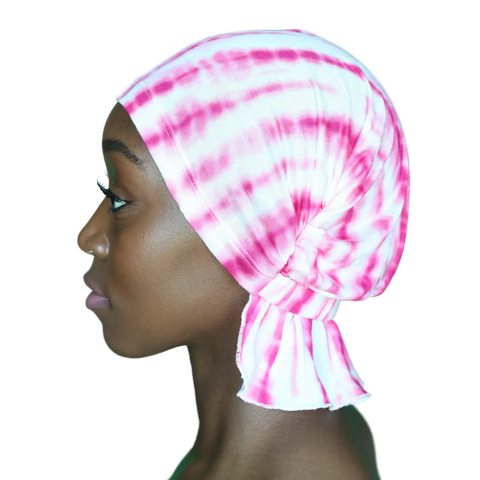 A woman in profile wears the TANISHA CHEMOBEANIE® by Chemo Beanies®, a pink and white tie-dye beanie covering her hair and tied at the back, set against a black background.