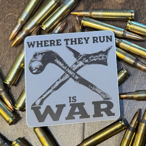 An American Lictor Sticker: Where They Run Is War, featuring crossed tomahawks and bold text, rests on a wooden surface among scattered rifle bullets.
