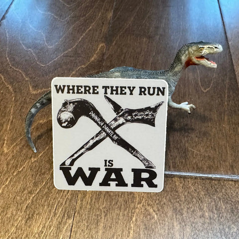 A toy dinosaur stands on wood behind the American Lictor Where They Run Is War sticker, featuring two crossed hammers above WAR, creating a fierce scene.