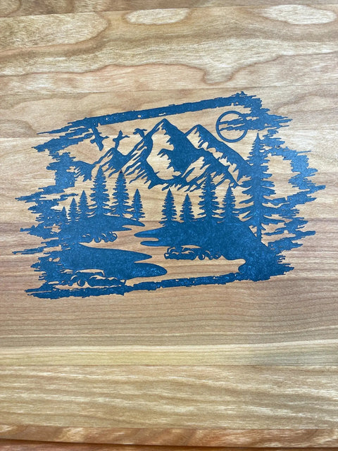 An intricately carved scenic mountain landscape with pine trees, river, and mountains is featured on the Blue Steel Epoxy Inlay Cherry Serving Tray With Grooved Outer Edge by B. James Design & Woodwork.