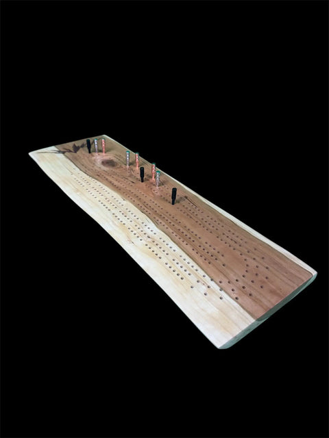 The Cherry Live Edge Cribbage Board With Mountain Engraving by B. James Design & Woodwork is shown with playing pegs in some holes, set against a black background.