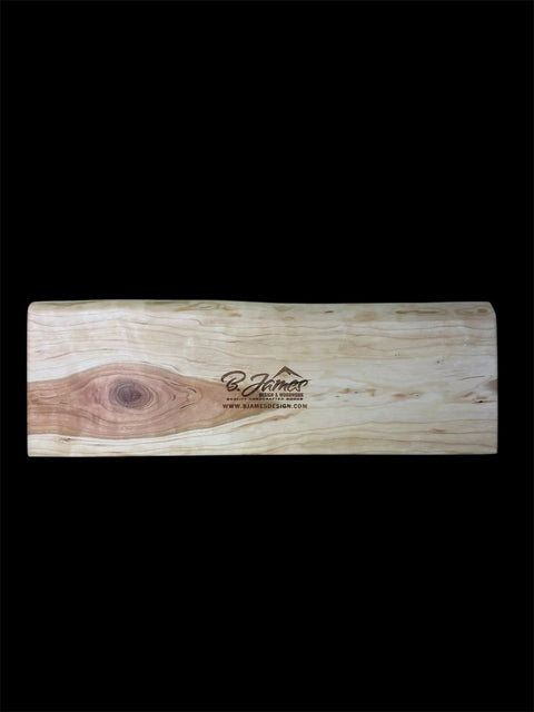 The Cherry Live Edge Cribbage Board with Mountain Engraving by B. James Design & Woodwork features natural cherry wood grain, a knot on the left, and center engravings of the brand logo, website, and a mountain, displayed on a black background.