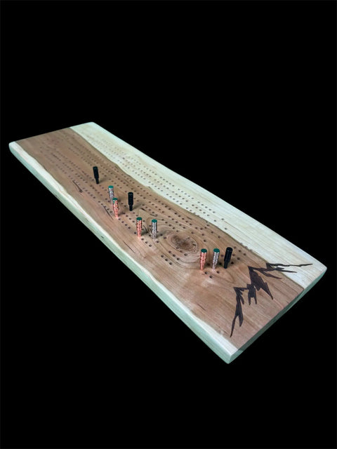 The Cherry Live Edge Cribbage Board With Mountain Engraving by B. James Design & Woodwork features pegs in various positions on a handcrafted board, with a mountain engraving on one corner, set against a black background.