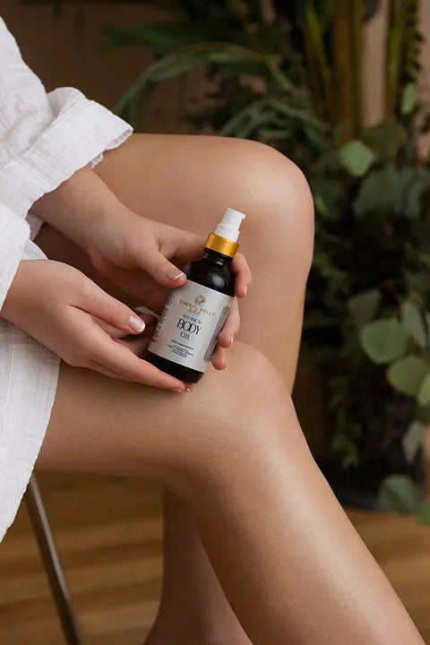 A person in a white robe sits cross-legged, holding a dark bottle of Cheryl Kelly & Co. Botanical Body Oil amid lush greenery and wooden floors, creating a serene vibe.