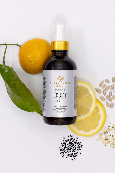 Cheryl Kelly & Co. Botanical Body Oil is displayed on a white background, surrounded by a whole lemon with leaf, a lemon slice, mixed seeds, and black seeds to highlight this luxurious product.
