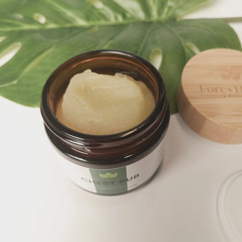 A jar of Forev'Herbs Chest Rub with creamy, pale yellow balm sits open on a white surface, accompanied by a green leaf and wooden lid in the background. The label displays “CHEST RUB.”.