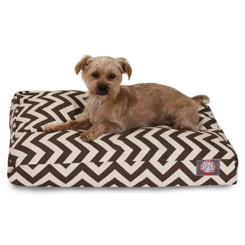 A small brown dog lies on the Majestic Pet Chevron Rectangle Dog Bed with a removable cover and front logo tag, looking directly at the camera. The bed features a stylish black and white chevron pattern.