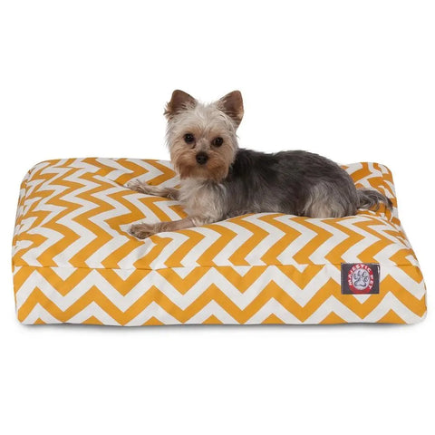 A small Yorkshire Terrier lies on the Majestic Pet Chevron Rectangle Dog Bed, featuring a yellow and white chevron pattern, red corner label, premium polyester fill for comfort, and a washable cover, looking directly at the camera.