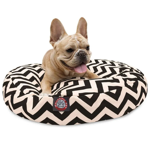 A tan French Bulldog with large ears relaxes on the Majestic Pet Chevron Round Dog Bed, featuring a black and white zigzag design. This washable dog bed keeps your pet cozy and makes cleanup simple. The dog looks content with its tongue out.