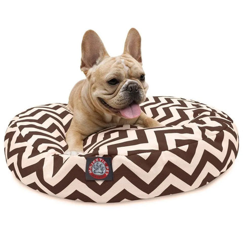 A light brown French Bulldog lies happily with its tongue out on the Majestic Pet Chevron Round Dog Bed, featuring a brown and white zigzag pattern and a washable, removable cover for easy cleaning.