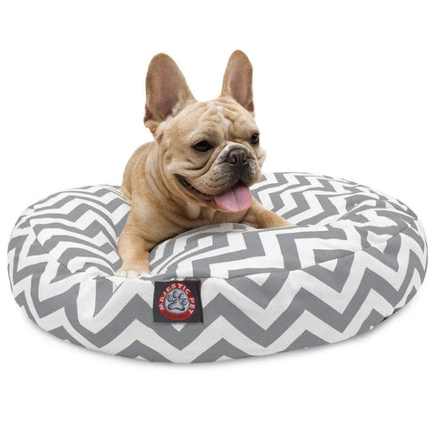 A light brown French Bulldog rests happily on the Majestic Pet Chevron Round Dog Bed, featuring a gray and white chevron pattern, red and gray brand badge, removable washable cover, and round design.