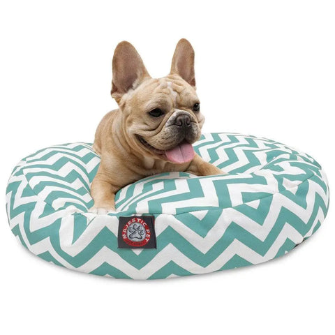 A light brown French Bulldog lounges happily on the Majestic Pet Chevron Round Dog Bed, featuring a teal and white chevron pattern and a red and white label on the front.