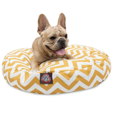 A French Bulldog rests on the Chevron Round Dog Bed by Majestic Pet, featuring a yellow and white chevron pattern, red logo on the front, and a removable washable cover for easy cleaning. The dog looks alert with its tongue out.