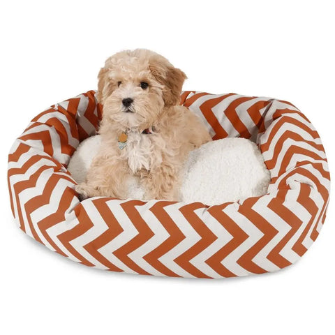 A small, fluffy light brown dog sits in the Majestic Pet Chevron Sherpa Bagel Dog Bed, featuring water-resistant outdoor polyester with a white and orange zigzag pattern and a plush white cushion inside.