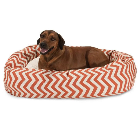 A brown dog relaxes in the Majestic Pet Chevron Sherpa Bagel Dog Bed, featuring an orange and white zigzag pattern and water-resistant fabric, facing the camera with its mouth slightly open.