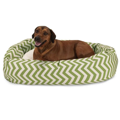 A brown dog lounges in the Majestic Pet Chevron Sherpa Bagel Dog Bed, featuring a green and white zigzag pattern and water-resistant outdoor-treated polyester, looking at the camera with its mouth slightly open.