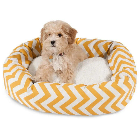 A small fluffy dog sits in the Majestic Pet Chevron Sherpa Bagel Dog Bed, featuring a yellow and white zigzag pattern, durable outdoor-treated polyester, and a soft white cushion for enhanced spine support.