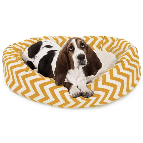 A basset hound with brown, black, and white fur lies in the Majestic Pet Chevron Sherpa Bagel Dog Bed, featuring water-resistant outdoor-treated polyester and a yellow and white chevron pattern, looking up with gentle eyes.