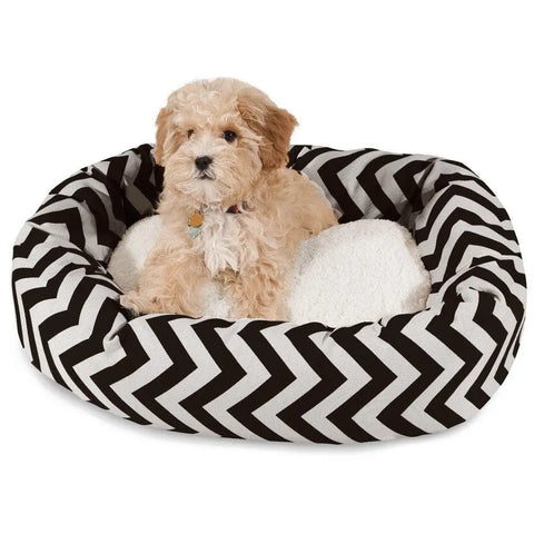 A small, fluffy light brown dog sits in the Majestic Pet Chevron Sherpa Bagel Dog Bed, which features a round black-and-white chevron pattern, soft white cushion, and durable outdoor-treated polyester.